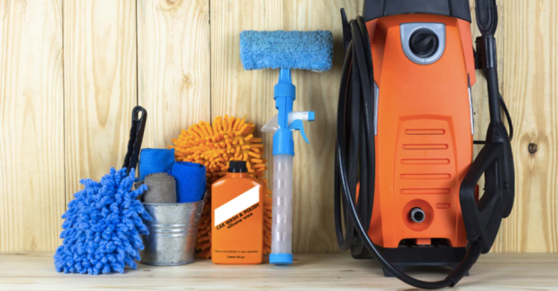 storing-pressure-washer Car cleaning equipment arranged on a wooden surface, including a blue mop, orange microfiber mitt, detergent bottle, metal bucket with cloths, spray bottle, and pressure washer.
