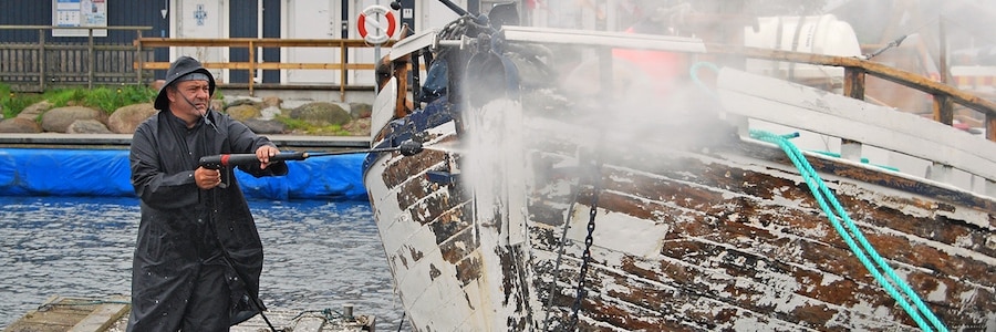 a fisherman on a wharf using a pressure washer to strip paint off of a boat