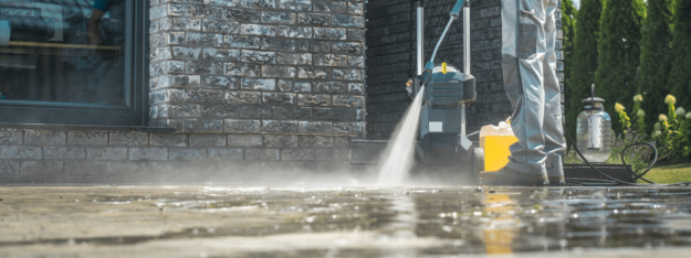 Common pressure washer problems Person pressure washing a patio near a brick building with a hose, spray bottle, and cleaning equipment nearby.
