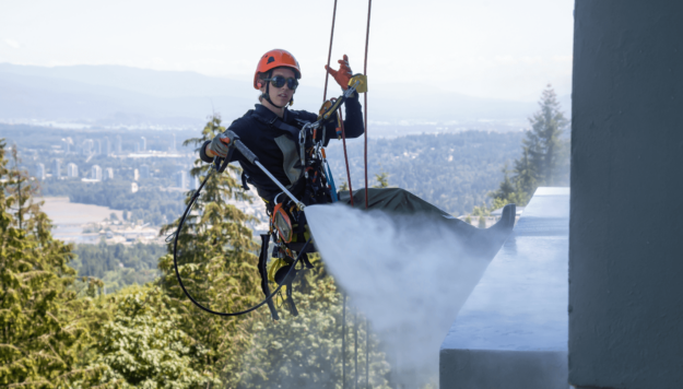 many applications of commercial pressure wshers (1) A worker wearing safety gear and a helmet is suspended on ropes while pressure washing a building facade with a city and forest visible in the background.