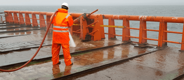 safely operate pressure washer (1) Person in an orange waterproof suit and white helmet using a hose to wash a wet ship deck in front of an orange railing with the ocean in the background.
