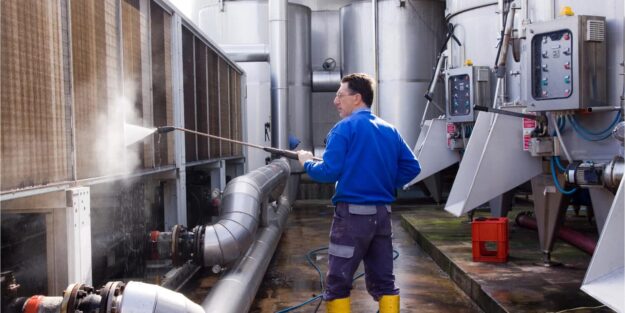 industrial pressure washing A man in a blue shirt and yellow boots uses a pressure washer on industrial equipment in a factory setting.