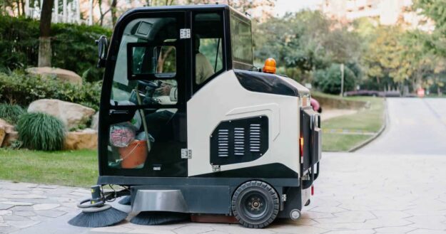 Using floor scrubber for concrete (1) A street sweeper vehicle on a paved path with trees and grass in the background.