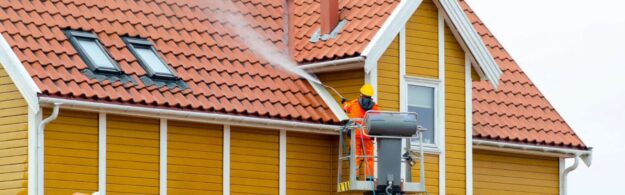 role of pressure washing exterior painting A worker in orange safety gear uses a power washer on the roof of a yellow house with red tiles.
