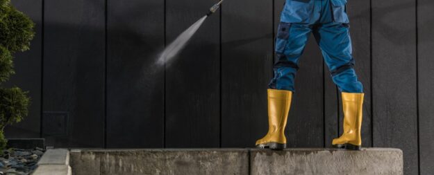 role of pressure washing landscaping Person wearing blue work clothes and yellow boots, power washing a dark wall.