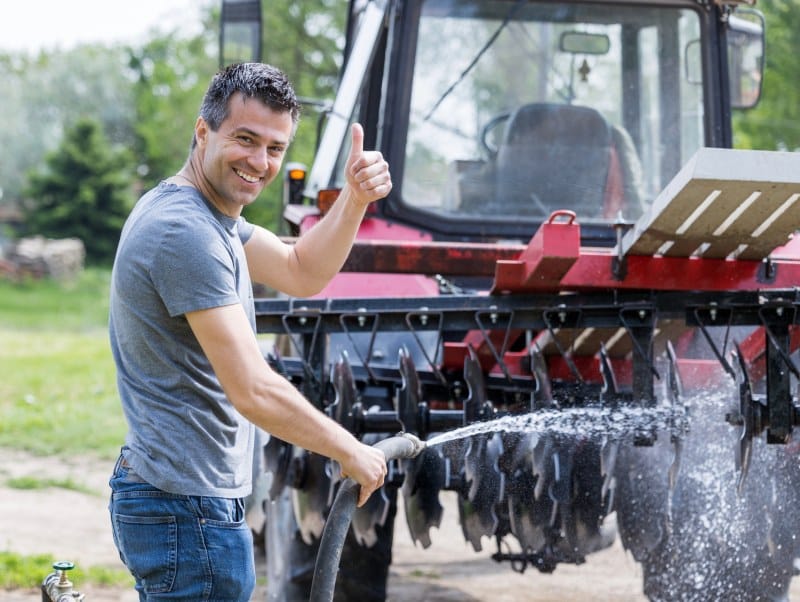 The Role of Pressure Washing in Agricultural Equipment Maintenance Farmer pressure washing a red tractor on his farm. He smiles at the camera with one thumb up in the air and the other holding the hose.