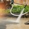 Man in long pants and black rubber boots pressure washing a concrete walkway.